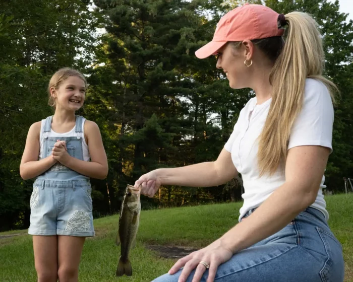 Adult holding a fish while smiling child stands nearby on grassy lakeshore