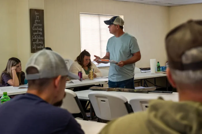 Instructor speaking to group in classroom with rifle on table during safety course