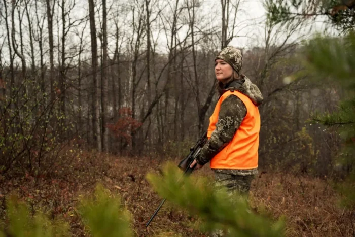 Person in camouflage and blaze orange vest holding rifle in wooded landscape