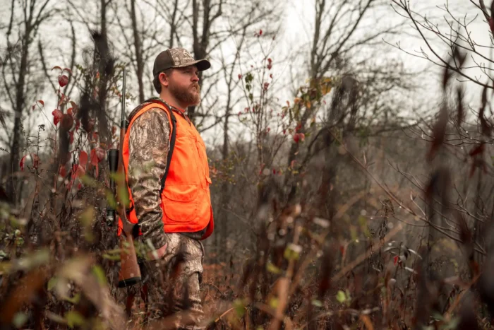 Person in camouflage and blaze orange vest standing in brush with rifle slung