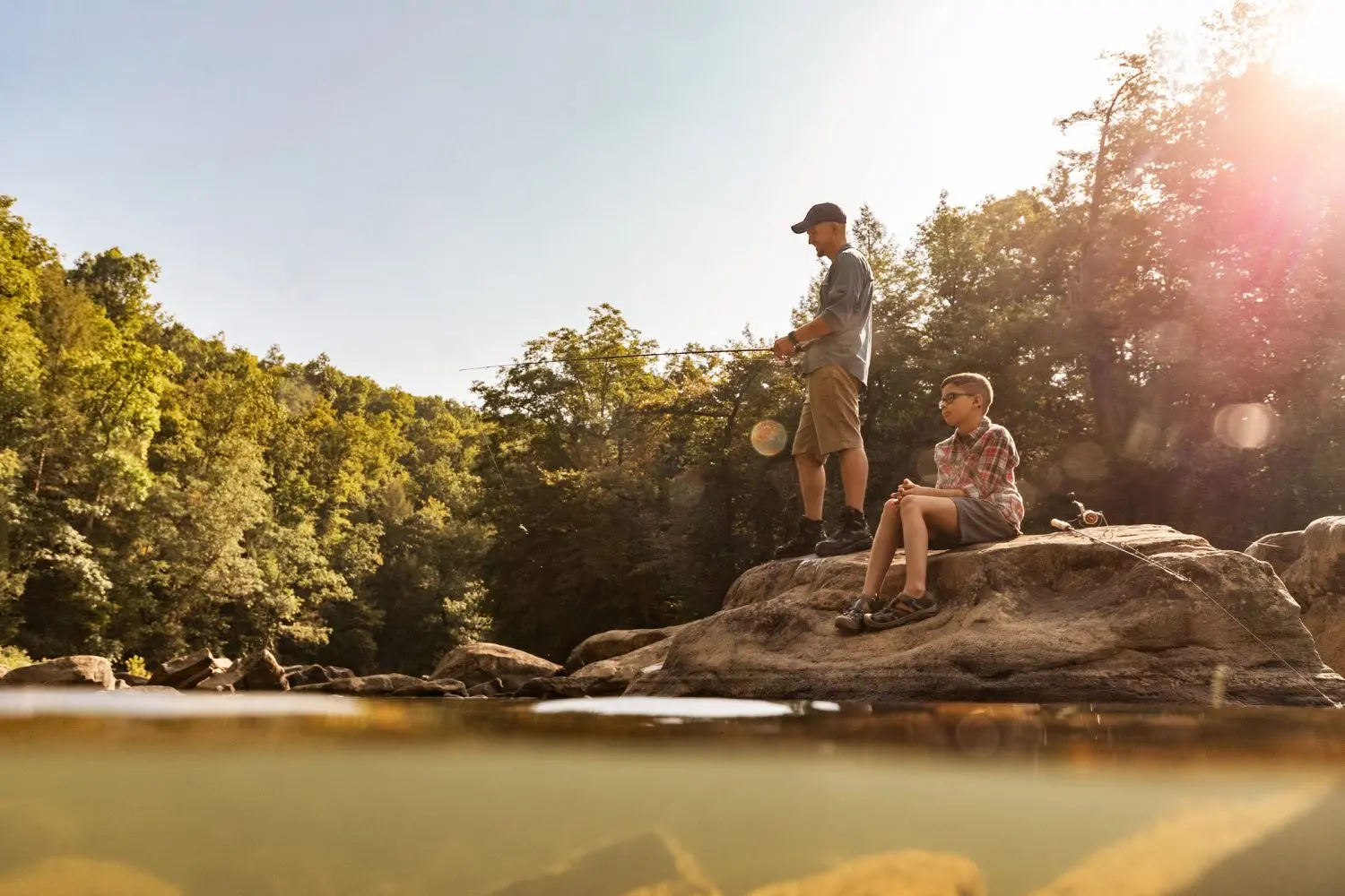 Adult fishing from a rock while a child sits beside a river at sunset