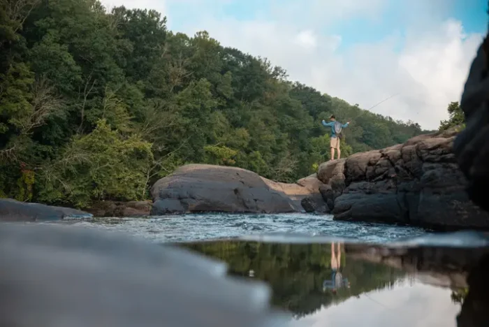 Person fishing from large rocks in a river with forested hills behind