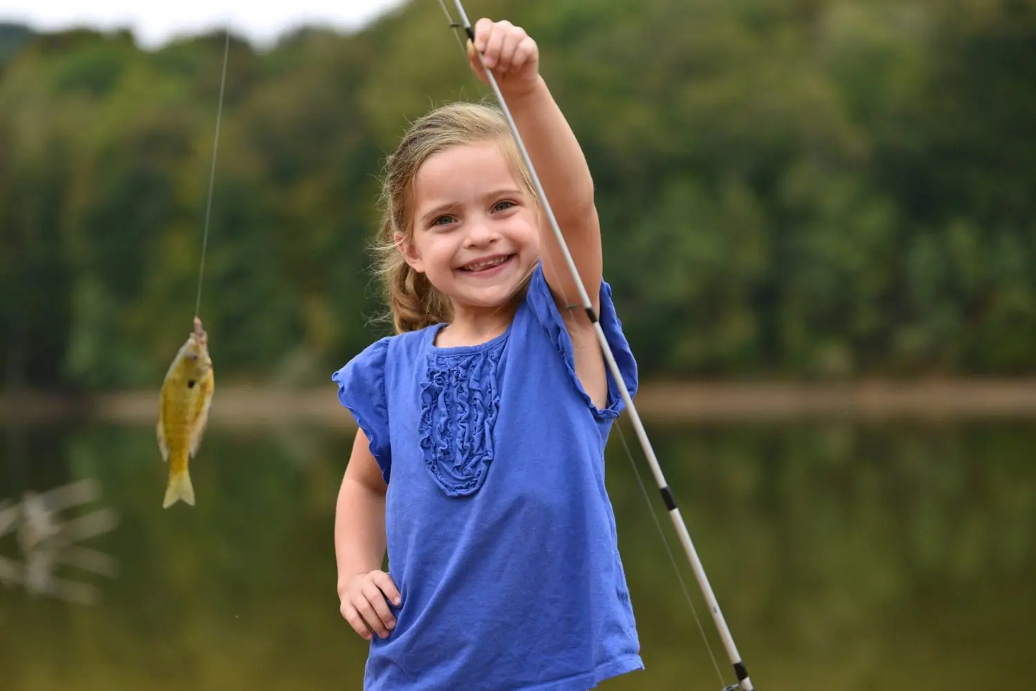 Child holding a small fish on a line while standing near a lake