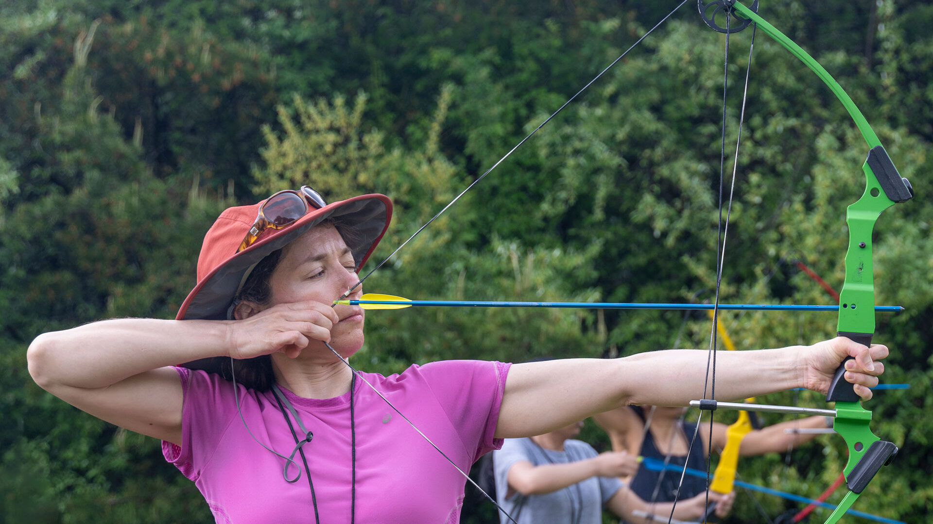 Woman in purple shirt draws back a bow