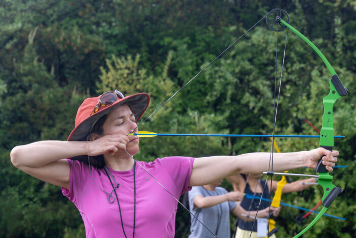 Woman in purple shirt draws back a bow